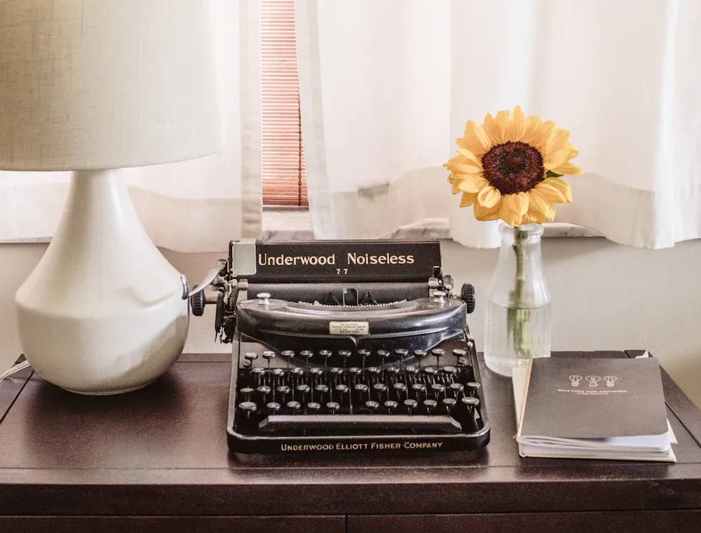A vintage typewriter on a desk beside a single sunflower