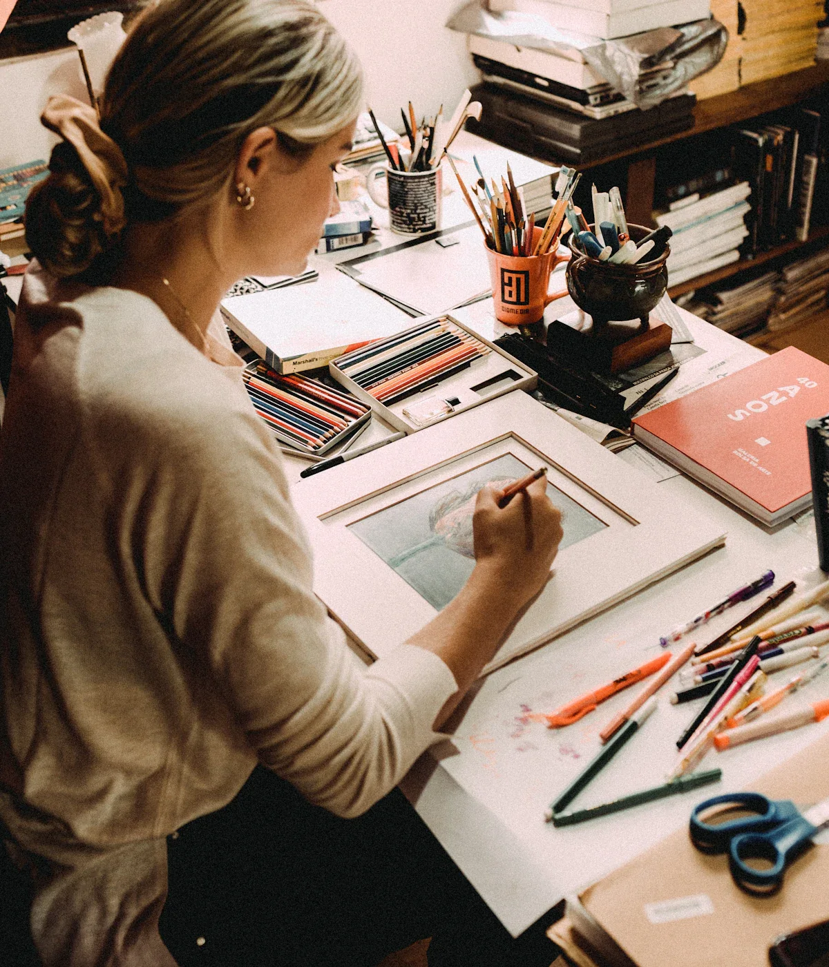 An illustrator drawing at a desk covered in pencils, paper and books