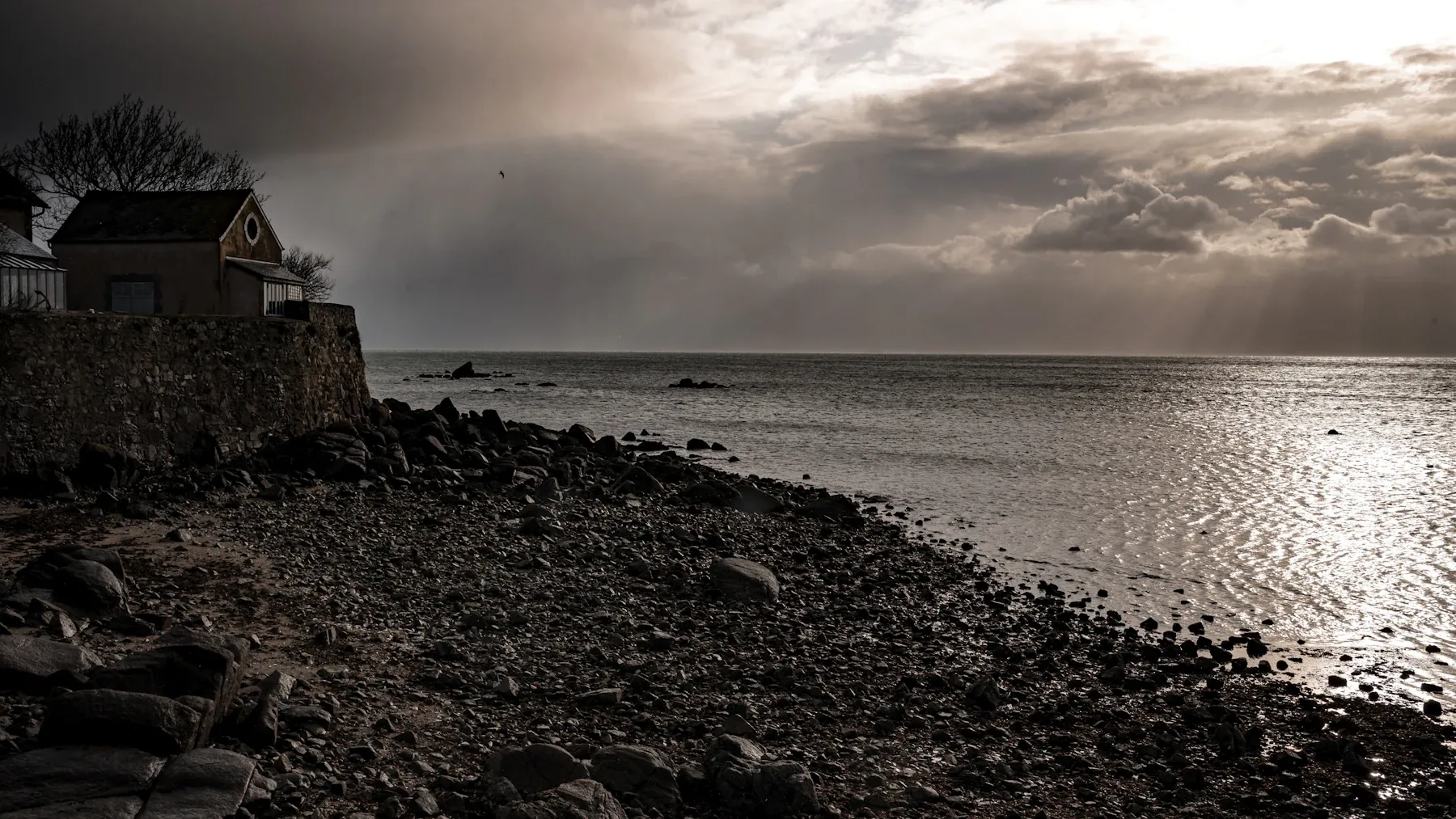 Film still: a stone cottage on a rocky shore as light breaks through storm clouds