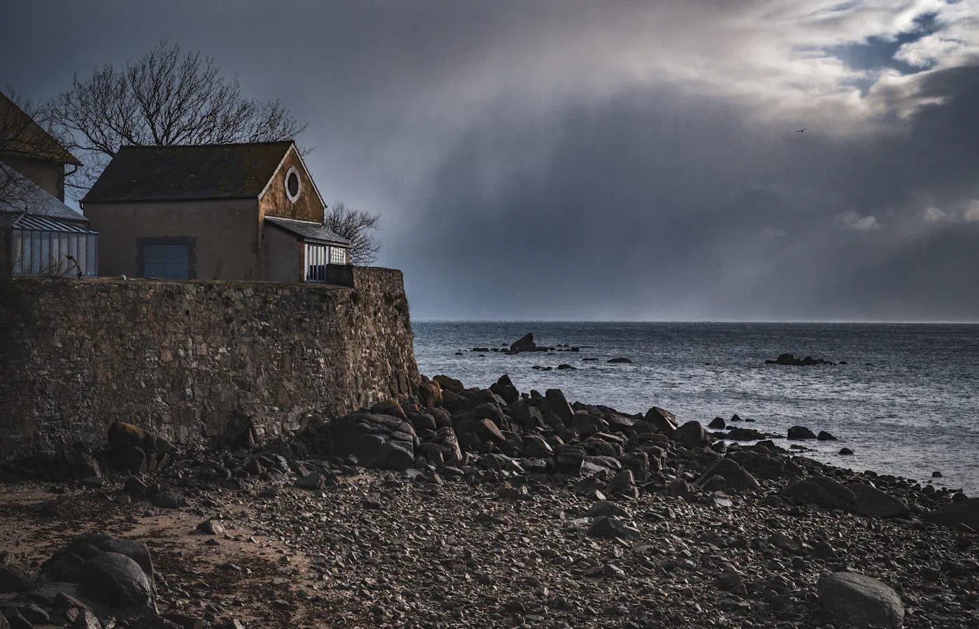 Film still: a weathered house on a sea wall under a storm sky
